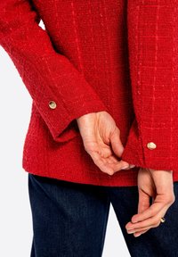 Giacca rossa con texture e un motivo intrecciato. Accenti di bottoni dorati sui polsini. Mani posizionate vicino all'orlo, che mettono in evidenza i dettagli delle maniche.