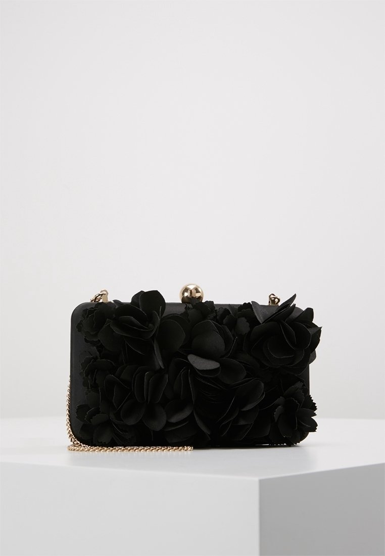 Clutch rettangolare nero adornato con applicazioni floreali testurizzate, tracolla in chain dorata e chiusura sferica dorata, posato su un piedistallo bianco.