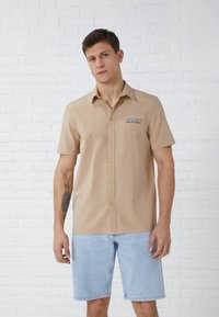 Short-sleeve, beige button-up shirt with a blue logo on the left chest. Worn with light blue denim shorts, against a white brick background.