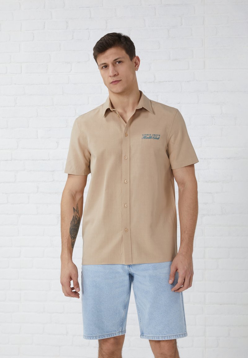 Short-sleeve, beige button-up shirt with a blue logo on the left chest. Worn with light blue denim shorts, against a white brick background.