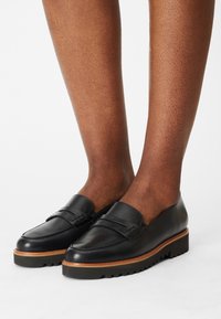Mocasines de cuero negro con un diseño cosido y un acento de madera sobre una suela gruesa negra, mostrados llevados en piernas con piel texturizada.