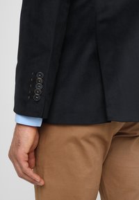 Mörk marinblå blazer med en texturerad tyg. Har kontrasterande ljusblå innerfoder vid ärmarna och sex knappar på ärmen med beige detaljer.