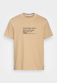 Beige bavlněné tričko s krátkým rukávem. Na přední straně je černý text: "Calvin Klein Jeans," adresa a PSČ.