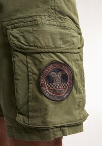 Olive green cargo shorts with a patch featuring an eagle and checkered pattern, multiple pockets, and a textured fabric.