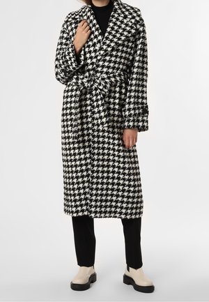 Personne portant un manteau ceinturé à motif pied-de-poule noir et blanc par-dessus un pantalon noir et des bottes à plateforme beige, debout sur un fond blanc.