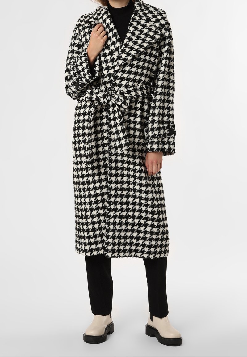 Personne portant un manteau ceinturé à motif pied-de-poule noir et blanc par-dessus un pantalon noir et des bottes à plateforme beige, debout sur un fond blanc.