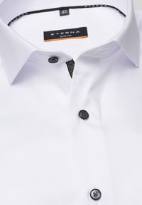 Witte slim-fit blouse van soepel materiaal. Heeft een zwarte knoopsluiting, zwarte knopen en een gestreepte accenten aan de binnenkant van de kraag.
