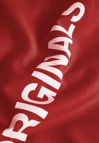 Red fabric featuring bold, white lettering of the word "ORIGINALS," with a smooth texture and slight sheen.
