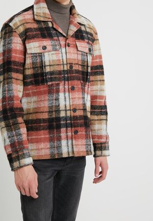 Mann trägt eine rote, beige, schwarze und weiße karierte Jacke mit Knöpfen über einem braunen Rollkragenpullover, kombiniert mit dunkelgrauen Jeans, vor weißem Hintergrund.