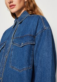 Jeans srajca v temno modri barvi z klasičnim ovratnikom, dvema žepoma na prsih in kontrastnimi šivi. Ohlapen kroj in mehak material.