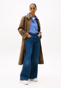 Brown trench coat over a blue striped shirt, paired with wide-leg blue jeans and white sneakers, displayed against a plain background.