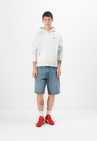 Jovem vestido com um hoodie cinza claro, calções de ganga até ao joelho, meias brancas e ténis vermelhos da Puma, de pé contra um fundo branco.