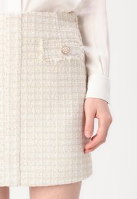 Beige tweed mini-jurk met een gestructureerd patroon, met een voorzak versierd met een decoratieve broche.