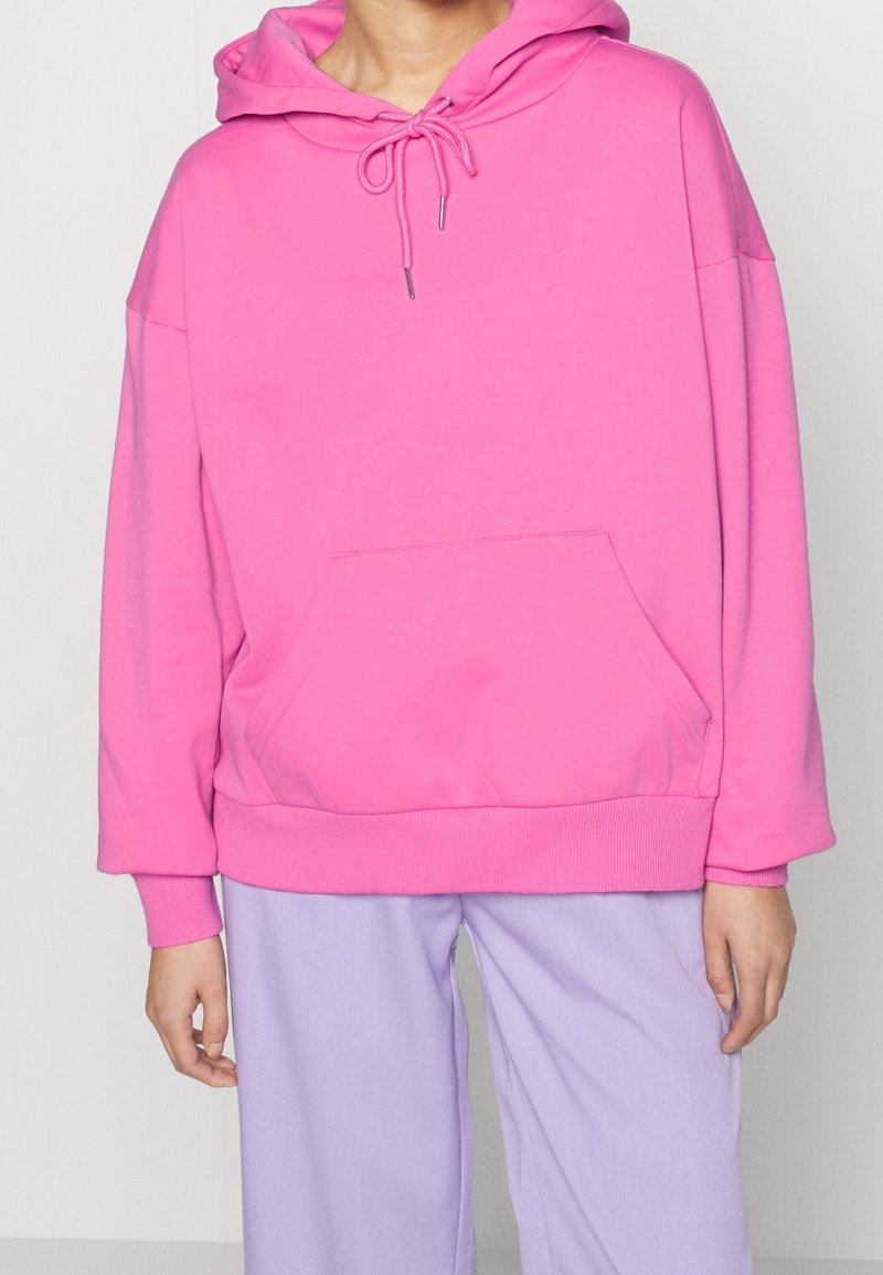 Personne portant un sweat à capuche oversize rose vif avec poche avant et pantalon ample lavande sur un fond uni.