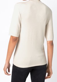 Osoba oblečená v béžovém pleteném tričku s krátkým rukávem a černých kalhotách, zobrazená zezadu na jednoduchém bílém pozadí.