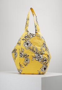 Borsa di tessuto gialla con una forma contornata, decorata con un motivo floreale nero e due spallacci per il trasporto.