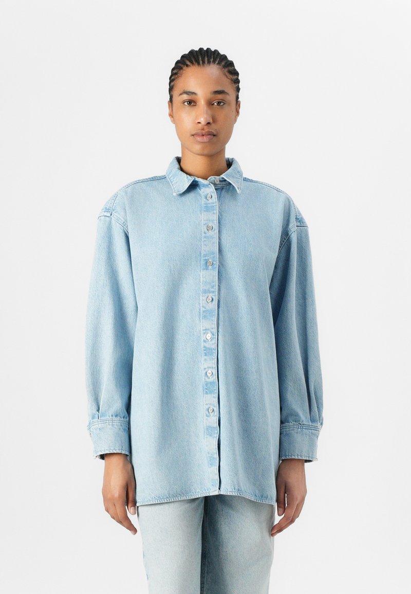 Lyseblå denim oversized skjorte med knappestolpe, klassisk krage og lange ermer. Har en løs passform og subtil tekstur.