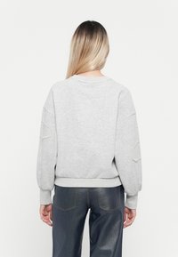 Kvinne med langt blondt hår iført en lys grå genser med perlepynt på ermene og mørk blå jeans, sett fra bak.