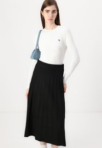 Hvit ribbestrikket langermet topp med liten logo, kombinert med et gulvlengde svart plisset skjørt. Modellen bærer en liten blå skulderveske.