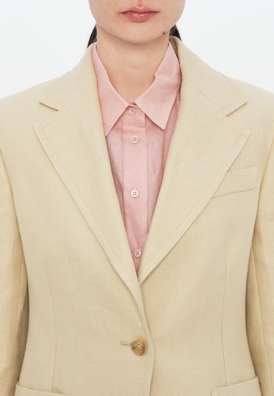 Beige herringbone blazer with a single button, notched lapels, and a front pocket, layered over a light pink button-up shirt.