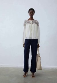 Weiße Bluse mit Spitzenüberlagerung und Pünktchen, kombiniert mit dunkelblauen Weitenhose. Das Modell trägt eine beigefarbene Tote-Bag und leichte Sandalen.