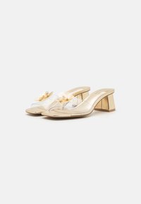 Mules à talon transparent en or avec une sangle transparente. Accents floraux décoratifs sur les sangles. Fini lisse et talon bloc structuré.