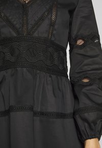 Robe noire avec des accents en dentelle texturée, broderie ondulée et manches bouffantes avec détails découpés. Composée d'un tissu léger et lisse.