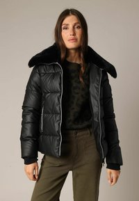 Zwarte donzen jas met een grote, nepbondkraag en witte ritssluitingen. Het model combineert dit met een gestructureerde groene trui en olijfgroene broek.