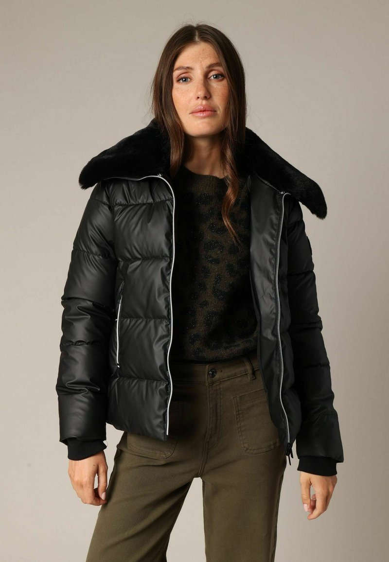 Zwarte donzen jas met een grote, nepbondkraag en witte ritssluitingen. Het model combineert dit met een gestructureerde groene trui en olijfgroene broek.