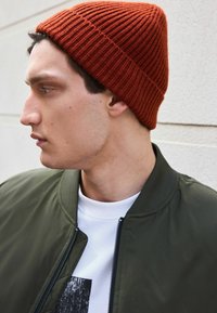 Rdzawy, dzianinowy beret o fakturze prążkowanej, noszony z oliwkowo-zieloną kurtką bombką oraz białą koszulką z czarną grafiką.