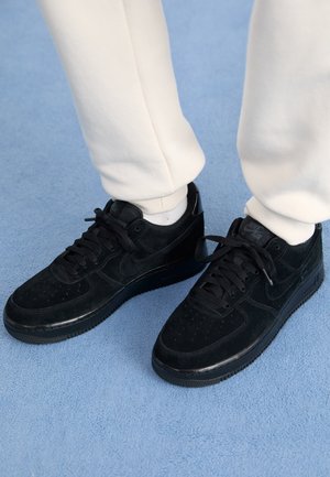 Personne portant des baskets Nike Air noires avec des chaussettes blanches et un pantalon couleur crème, debout sur un tapis bleu clair.