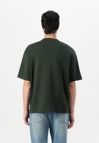 T-shirt en coton vert foncé avec une coupe décontractée et des manches courtes, présentant un col rond et un ourlet droit. Assorti à un jean bleu clair.