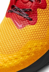 Sneaker Nike con tomaia in mesh gialla e rossa, materiali testurizzati, accenti rosa e lacci neri su una suola scura.