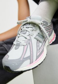 Primo piano di una sneaker New Balance grigia e rosa con lacci bianchi, indossata con calze bianche, poggiata su una superficie grigia chiara.