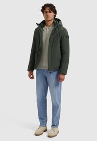 Donkergroene pufferjas met een capuchon, ritssluiting en zijzakken, gedragen over een lichtgrijze polo, gecombineerd met lichtblauwe jeans en beige schoenen.