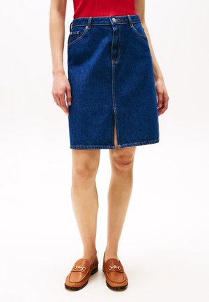 Femme portant une jupe en jean bleue longueur genou avec une fente à l'avant, des mocassins marron et un haut sans manches rouge, debout devant un fond blanc.