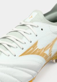 Hvit fotballsko med lysegrå detaljer, med et teksturert design, gullfarget logo og standard snøringssystem. Egnet for utendørs bruk.