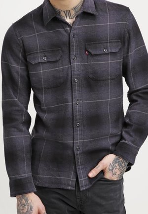 Homme portant une chemise à carreaux grise foncée boutonnée avec deux poches poitrine, montrant des tatouages sur le cou et les mains, une main dans la poche.