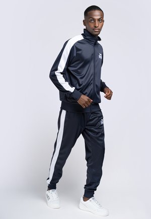 Tuta della marina realizzato in materiale lucido con strisce laterali bianche. Include una giacca con zip e pantaloni con elastico in vita, completati da sneakers bianche.