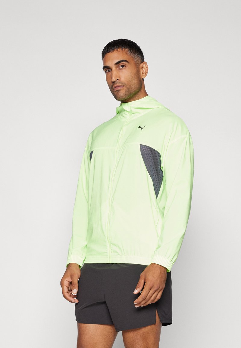 Homme portant une veste zippée Puma vert fluo avec des accents en maille grise et un short de sport noir, prenant la pose devant un fond clair.