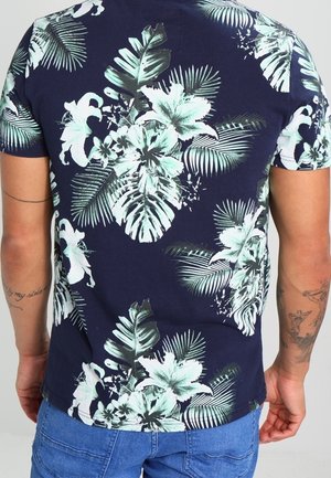 Mann trägt ein marineblaues T-Shirt mit grünen und weißen tropischen Blumen- und Palmenblattmustern, kombiniert mit blauen Jeansshorts.