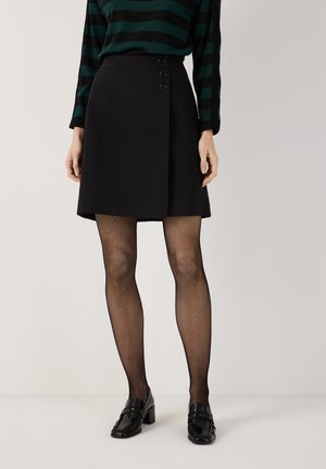 Frau trägt einen schwarzen, mit Knöpfen versehenen Wickelrock, schwarze Netzstrumpfhosen, schwarze Loafers mit Absatz und ein dunkelgrünes und schwarzes gestreiftes Oberteil.