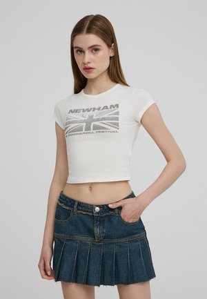 Maglietta bianca cropped con stampa grafica grigia che presenta "NEWHAM UK ROCK & ROLL FESTIVAL" e un Union Jack. Abbinata a una gonna in denim plissettata.