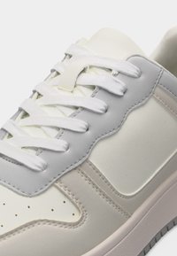 Gros plan sur une chaussure de sport blanche et grise avec des lacets blancs, des coutures visibles et des perforations sur la zone des orteils.