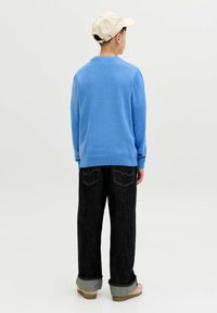 Garçon portant une casquette beige, un pull bleu clair, un jean foncé retroussé et des chaussures beiges, debout de dos sur un fond blanc uni.