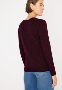 Maglione bordeaux a maniche lunghe, lavorato a maglia con sottili striature rosse, collo arrotondato e silhouette aderente. Abbinato a jeans a gamba larga di un blu chiaro.