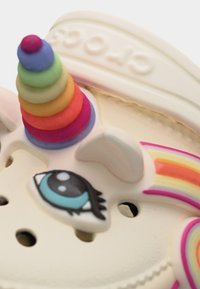 Gros plan sur une chaussure crème avec une corne de licorne empilée colorée, un œil de dessin animé bleu, et un détail arc-en-ciel sur le côté.