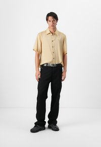 Camisa de manga corta, color amarillo claro, con botones y un logo en el pecho, combinada con pantalones negros y un cinturón negro con hebilla plateada.