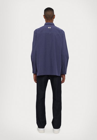 Camicia a quadri blu con orlo curvo, vestibilità comoda e maniche larghe, abbinata a pantaloni in denim scuro e sneakers bianche.