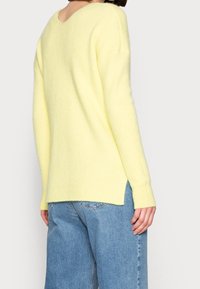 Person som bär en ljusgul stickad tröja med V-rygg och sidofickor, tillsammans med medelblå denimjeans.